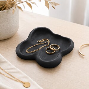 May include: A matte black, four-lobed, cloud-shaped trinket dish holding a thin gold chain and two gold rings. The dish has a smooth, rounded edge and a concave center, providing a functional space for small jewelry items and accessories.