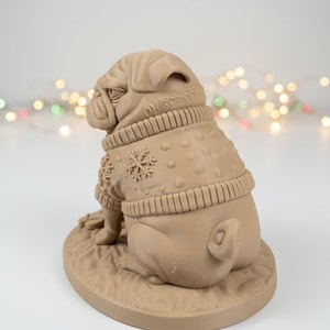Puede incluir: Una figura monocromática de color marrón claro impresa en 3D de un perro pug sentado sobre una base ovalada. El pug lleva un suéter texturizado con un patrón de copo de nieve en relieve en la espalda, cuello acanalado y dobladillo acanalado.