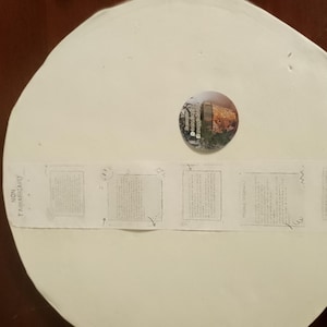 May include: A large, off-white, circular object with a small, round, colorful image in the center. A strip of paper with text and small square images is taped across the object. The object is on a dark brown surface.