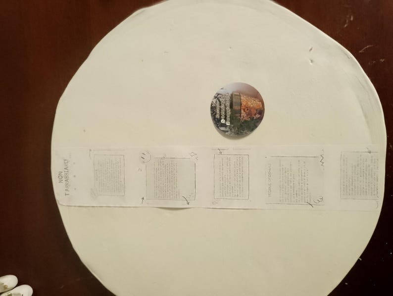 May include: A large, off-white, circular object with a small, round, colorful image in the center. A strip of paper with text and small square images is taped across the object. The object is on a dark brown surface.