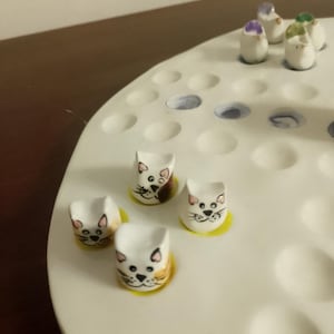 May include: A white ceramic game board with multiple holes, featuring small, cat-shaped game pieces with painted faces and yellow bases. Additional pieces with purple and green accents are also present.