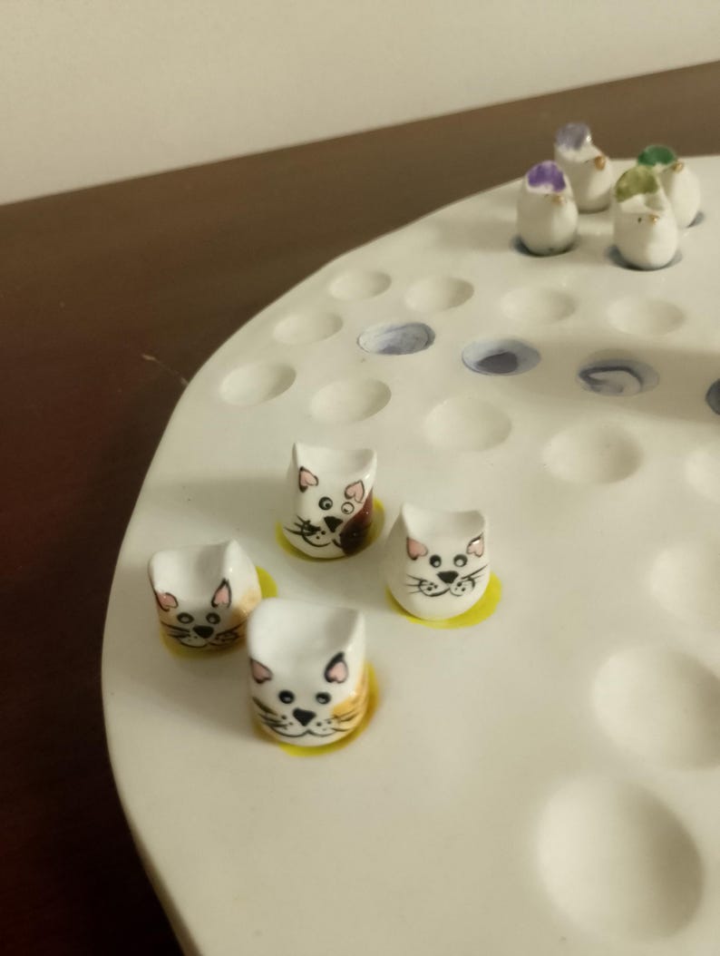 May include: A white ceramic game board with multiple holes, featuring small, cat-shaped game pieces with painted faces and yellow bases. Additional pieces with purple and green accents are also present.