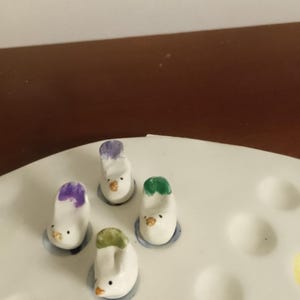 May include: A collection of four small, white ceramic bird figurines with colorful accents. Each bird has a different colored head, including purple, green, and white. They are placed on a white ceramic tray with circular indentations.