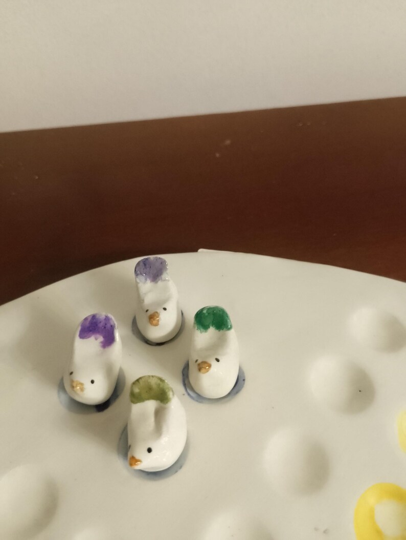 May include: A collection of four small, white ceramic bird figurines with colorful accents. Each bird has a different colored head, including purple, green, and white. They are placed on a white ceramic tray with circular indentations.