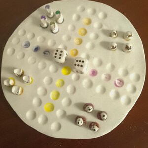 May include: A white ceramic board game with various game pieces. The game pieces include small, painted figures, dice, and colorful dots. The board has a circular shape with evenly spaced holes for the game pieces.