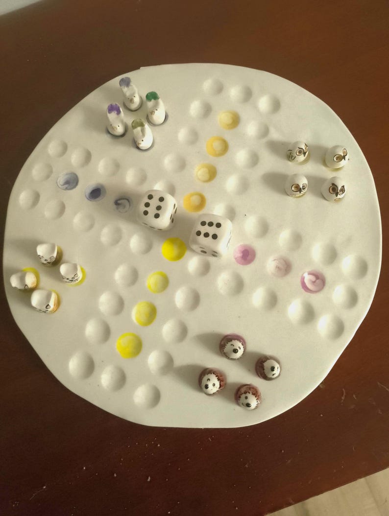 May include: A white ceramic board game with various game pieces. The game pieces include small, painted figures, dice, and colorful dots. The board has a circular shape with evenly spaced holes for the game pieces.