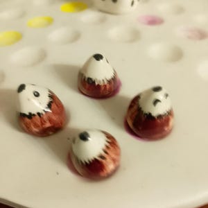 May include: Four ceramic hedgehog game pieces with white bellies, brown backs, and black eyes are arranged on a white game board. Two white dice with black dots are also present.