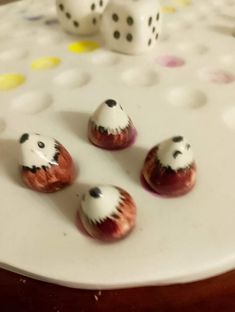 May include: Four ceramic hedgehog game pieces with white bellies, brown backs, and black eyes are arranged on a white game board. Two white dice with black dots are also present.