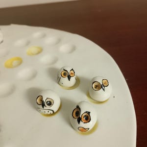 May include: Four small, white, owl-shaped ceramic figurines with orange and black details. The owls are arranged on a white, oval-shaped surface with yellow accents. The figurines have a hand-painted appearance.