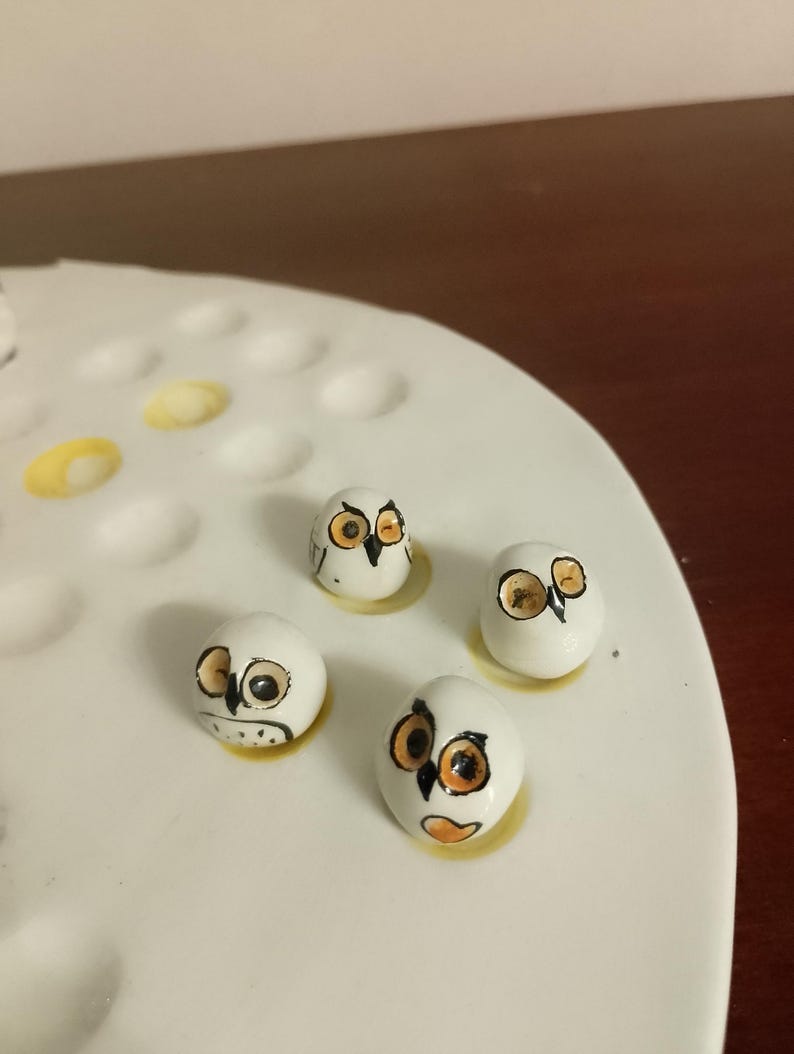 May include: Four small, white, owl-shaped ceramic figurines with orange and black details. The owls are arranged on a white, oval-shaped surface with yellow accents. The figurines have a hand-painted appearance.