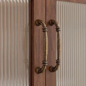 May include: A pair of antique brass-finished metal cabinet handles mounted on dark brown wooden cabinet doors. The doors feature vertical fluted glass panels set within the wood frame, creating a textured and layered appearance.