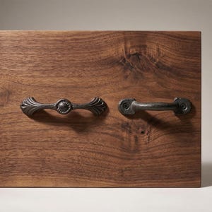 May include: Two antique-style drawer pulls on a wooden surface. One pull has a decorative, symmetrical design with a central circular element and curved ends. The other is a simple, rectangular handle. Both are dark grey.