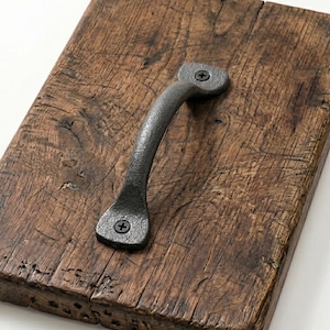 May include: A weathered wooden board with a dark, textured surface. A curved, dark grey metal handle is fixed to the wood with screws. The handle's design suggests it's for a drawer or cabinet, adding a touch of rustic charm.