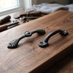 May include: Two black metal cabinet handles. One features a floral design with a central circular element and curved leaf-like details. The other is a simple, arched handle. Both are fixed with screws and rest on a wooden surface.