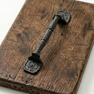 May include: A rustic, rectangular wooden serving board with a decorative, black metal handle. The handle features an ornate design and is attached with screws. The wood has a natural, weathered appearance.
