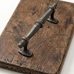 May include: A rectangular wooden serving board with a dark, textured, cast iron handle. The handle is attached with screws and has decorative detailing. The wood has a natural, aged appearance with visible grain and knots.