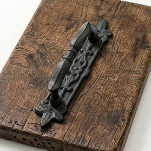 Può includere: Una maniglia nera e ornata, con maniglia e piastra posteriore decorative, montata su una tavola di legno rustica e invecchiata. La maniglia presenta un'intricata lavorazione a volute ed è fissata con viti.