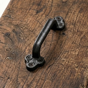 Könnte beinhalten: Ein schwarzer Gusseisen-Griff mit geschwungenem Design. Der Griff ist mit Schrauben an einer Holzoberfläche befestigt. Die Hardware hat eine rustikale, Vintage-Ästhetik, geeignet für Möbel oder Schränke.