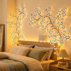 Op de afbeelding: Slaapkamerinterieur met een bed en kussens. De muur is versierd met verlichte bloemen takken wandkunst met blauwe bloemen. Een nachtkastje met een lamp, boeken en een mok is zichtbaar. Warme, uitnodigende sfeer.