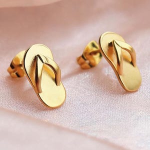 May include: A pair of gold-toned stud earrings shaped like miniature flip-flops. Each earring features a smooth, flat sole and a raised thong strap detail, finished with a polished metallic surface that reflects light across the small, detailed forms.