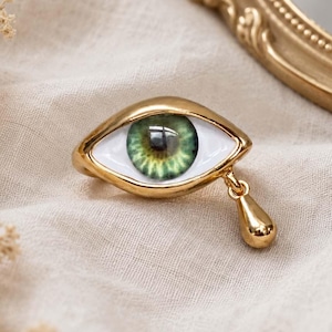 Puede incluir: Un broche dorado con forma de ojo, con esclerótica de esmalte blanco e iris verde realista. Un pequeño dije dorado en forma de lágrima cuelga de la esquina inferior del ojo, añadiendo un elemento colgante al diseño general.