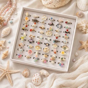 Könnte beinhalten: Eine weiße, rechteckige Präsentationsbox mit einer Sammlung von fünfzig Ringen. Jeder Ring ist mit einzigartigen dekorativen Elementen wie Miniatur-Muscheln, kleinen Perlen, bunten Kügelchen, floralen Akzenten und Metallteilen versehen.