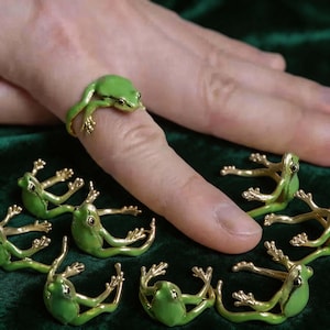 May include: Green frog rings with gold-colored accents. One ring is worn on a finger, with several others displayed nearby. The rings feature a detailed frog design, with the frog's body and legs wrapping around the finger.