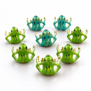 May include: A collection of eight small frog-shaped metal charms with gold-toned edges and enamel finishes in shades of bright green and teal. Each frog is depicted in a seated position with arms resting on its hips, featuring black painted eyes.