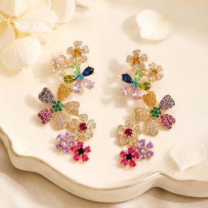 May include: A pair of floral earrings featuring multiple connected flower shapes encrusted with multicolored crystals. The petals display vibrant shades of pink, purple, blue, green, yellow, and clear stones, creating a cluster design that cascades downward in a bright, multi-colored arrangement.