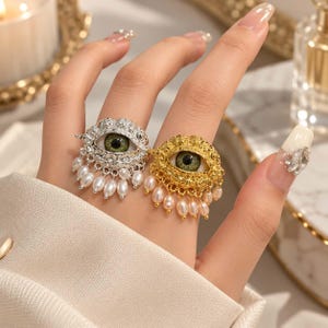 May include: Two rings featuring a realistic green eye design set within a textured metal frame. One ring is silver-toned and the other is gold-toned. Both rings are adorned with small, dangling white pearl-like beads along the bottom edge.