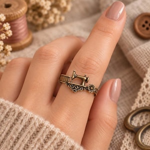 May include: A bronze-toned metal ring featuring a miniature sewing machine design integrated into the band. The band includes a small engraved measuring tape detail and a decorative floral accent, creating a cohesive, detailed accessory.