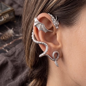 May include: A silver-toned ear cuff featuring a dragon head at the top, a wing extending across the upper ear cartilage, and a long, textured snake-like body that curves down along the outer edge of the ear, ending in a coiled tail at the lobe.