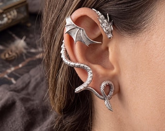May include: A silver-toned ear cuff featuring a dragon head at the top, a wing extending across the upper ear cartilage, and a long, textured snake-like body that curves down along the outer edge of the ear, ending in a coiled tail at the lobe.