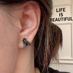 May include: A dark metallic fly-shaped stud earring worn on an earlobe. The insect design features detailed wings and a segmented body. In the upper right corner, the text LIFE IS BEAUTIFUL is printed in black capital letters against a white background.