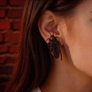 May include: A dark brown, insect-shaped earring featuring a detailed, textured body with prominent wings and two long, thin antennae extending upward. The piece is designed to sit within the ear, mimicking the appearance of a cockroach.