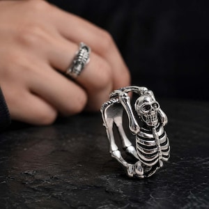 May include: A silver-toned ring featuring a detailed skeleton figure that wraps around the band. The skeleton has a visible skull, rib cage, and arm bones forming the structure of the ring, with dark oxidized accents highlighting the skeletal details.