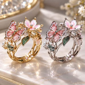 May include: Two rings featuring a butterfly and flower design with pink enamel wings, green enamel leaves, and small clear stones. One ring is gold-toned metal, and the other is silver-toned metal, both showcasing intricate floral and insect motifs.
