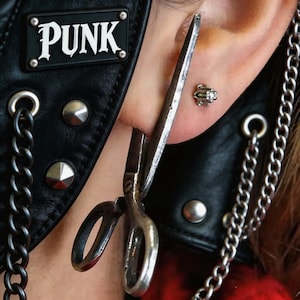 May include: A silver-toned earring shaped like a pair of scissors worn in an earlobe, accompanied by a small stud earring with a clear stone. The ear is positioned near a black leather garment featuring a metal plate with the text PUNK.