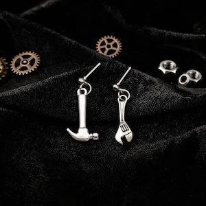Puede incluir: Pendientes plateados con forma de martillo y llave inglesa sobre tela negra. Los pendientes presentan un diseño de martillo y llave inglesa. Pequeños engranajes y tuercas metálicas están dispersos alrededor de los pendientes.