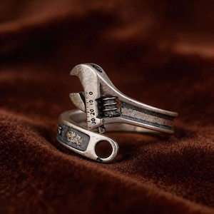 May include: A silver-colored adjustable wrench ring. The ring is designed to wrap around the finger, with the wrench head and handle forming the top and bottom. The wrench has a scale with numbers and a small decorative element.