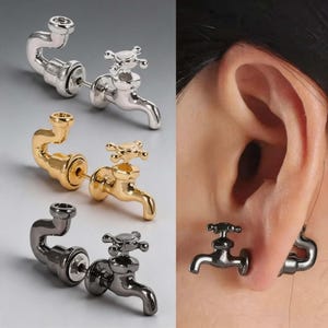 May include: A set of three stud earrings shaped like miniature water faucets in silver, gold, and black metal tones. Each earring features a cross-handle design and a curved spout, with one black faucet earring shown inserted into an earlobe.