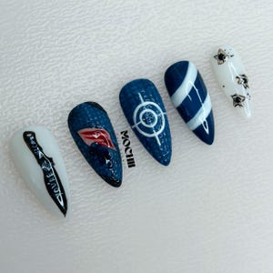 May include: Five almond-shaped press-on nails featuring varied designs: a white nail with a black knife and the text 'YOU'RE WELCOME', a textured dark blue nail with a red lip graphic, a textured blue nail with a white target, a blue and white striped nail, and a white nail with black star accents.