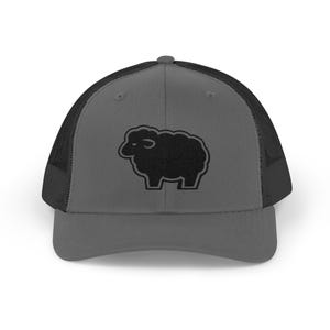 May include: A charcoal gray trucker hat featuring a black mesh back and a solid front panel. Centered on the front is an embroidered black sheep silhouette with a thick black outline, providing a distinct contrast against the gray fabric background.