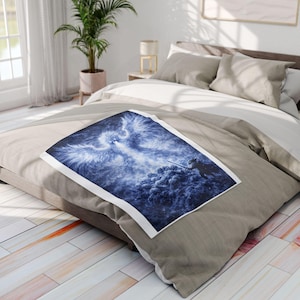Könnte beinhalten: Eine blau-weiße Decke mit einem Phönix-Design. Die Decke liegt auf einem Bett mit neutralfarbener Bettwäsche. Der Phönix wird mit ausgebreiteten Flügeln dargestellt, die ein helles Licht aussenden. Die Decke besteht aus weichem Material.