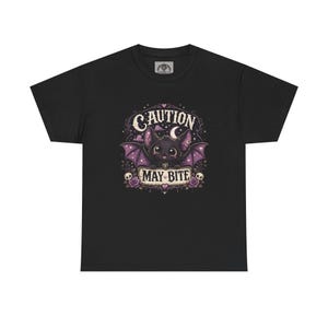 以下が含まれることがあります： 黒いTシャツ。漫画のコウモリのデザイン。コウモリは紫色の翼と「CAUTION MAY BITE」の文字があります。デザインにはバラ、頭蓋骨、月が含まれています。カジュアルウェアです。