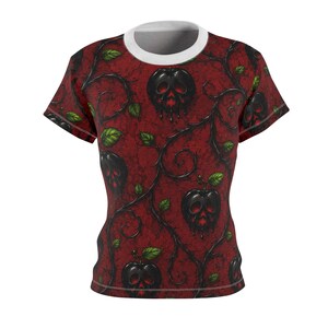 Può includere: T-shirt a maniche corte con girocollo bianco e sfondo rosso. La maglietta presenta un motivo ripetuto di mele nere con facce a forma di teschio, viti e foglie verdi. Il design ha un tema gotico o fantasy.