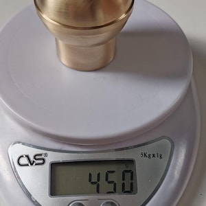 May include: A brass-coloured, spherical knob with a cylindrical base, resting on a digital scale. The scale shows a weight of 450 grams. The knob has a smooth, polished surface.
