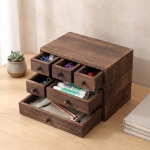 Könnte beinhalten: Ein dunkelbrauner Schreibtisch-Organizer aus Holz mit mehreren kleinen Schubladen. Die Schubladen sind geöffnet und zeigen verschiedene Büroartikel. Der Organizer ist gestapelt, mit einer Sukkulentenpflanze in einem kleinen Topf im Hintergrund.
