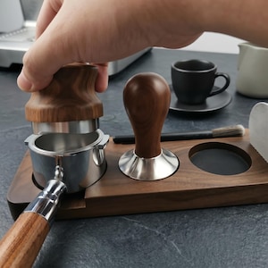 May include: A wooden espresso tamping station with a portafilter, tamper, and other coffee accessories. The tamper has a wooden handle and a stainless steel base. A hand is pressing down on the tamper. A black coffee cup and a small brush are also visible.