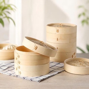 May include: A set of light-coloured bamboo steamer baskets with woven lids and stitched side details. The stack includes multiple cylindrical tiers, with one lid placed at an angle and another separate lid resting on the side, showing natural wood grain.